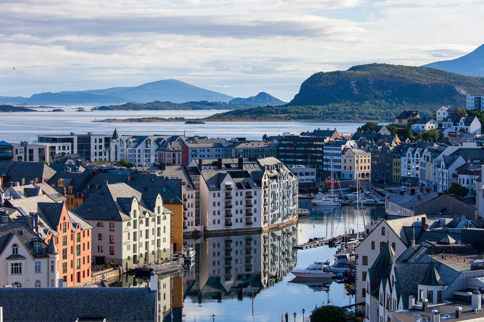 View of Ålesund’s colourful waterfront and harbour with reflections on calm water, surrounded by mountains and islands along Norway’s western coast.
