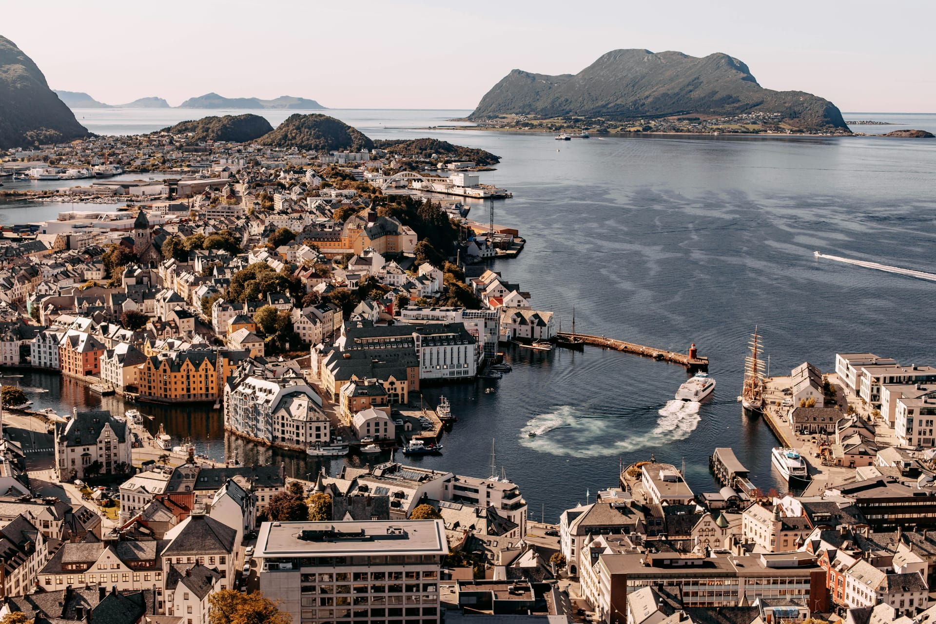 Aerial view of Ålesund, Norway, showcasing its colourful Art Nouveau buildings, harbour, and surrounding islands under a clear sky, with boats navigating through calm coastal waters.