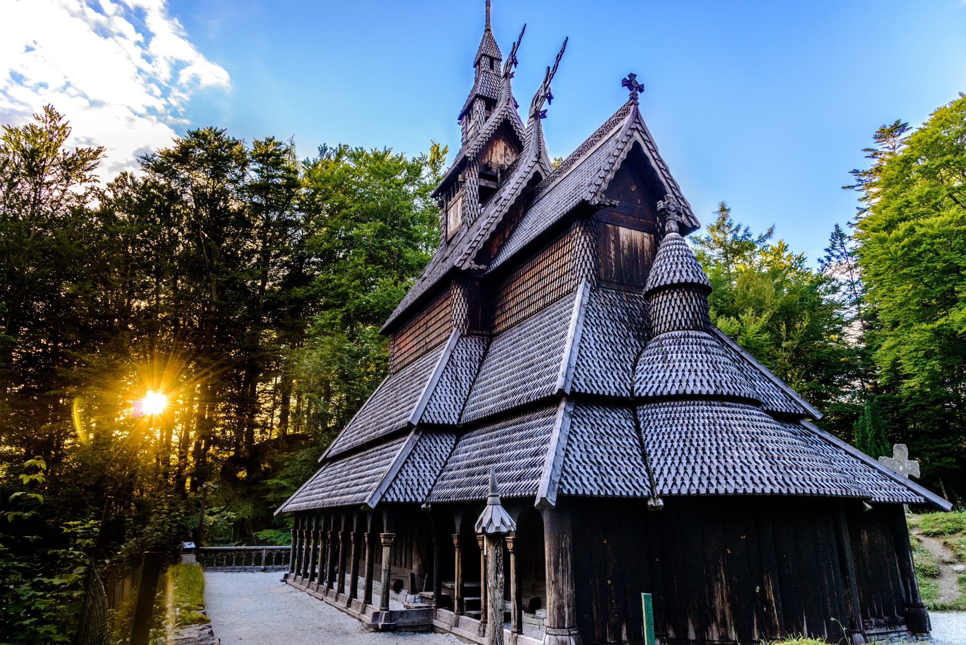 Fantoft Stave Church in Bergen, Norway, captured at sunset with warm sunlight filtering through the surrounding forest and highlighting the intricate wooden architecture of the medieval-style church.