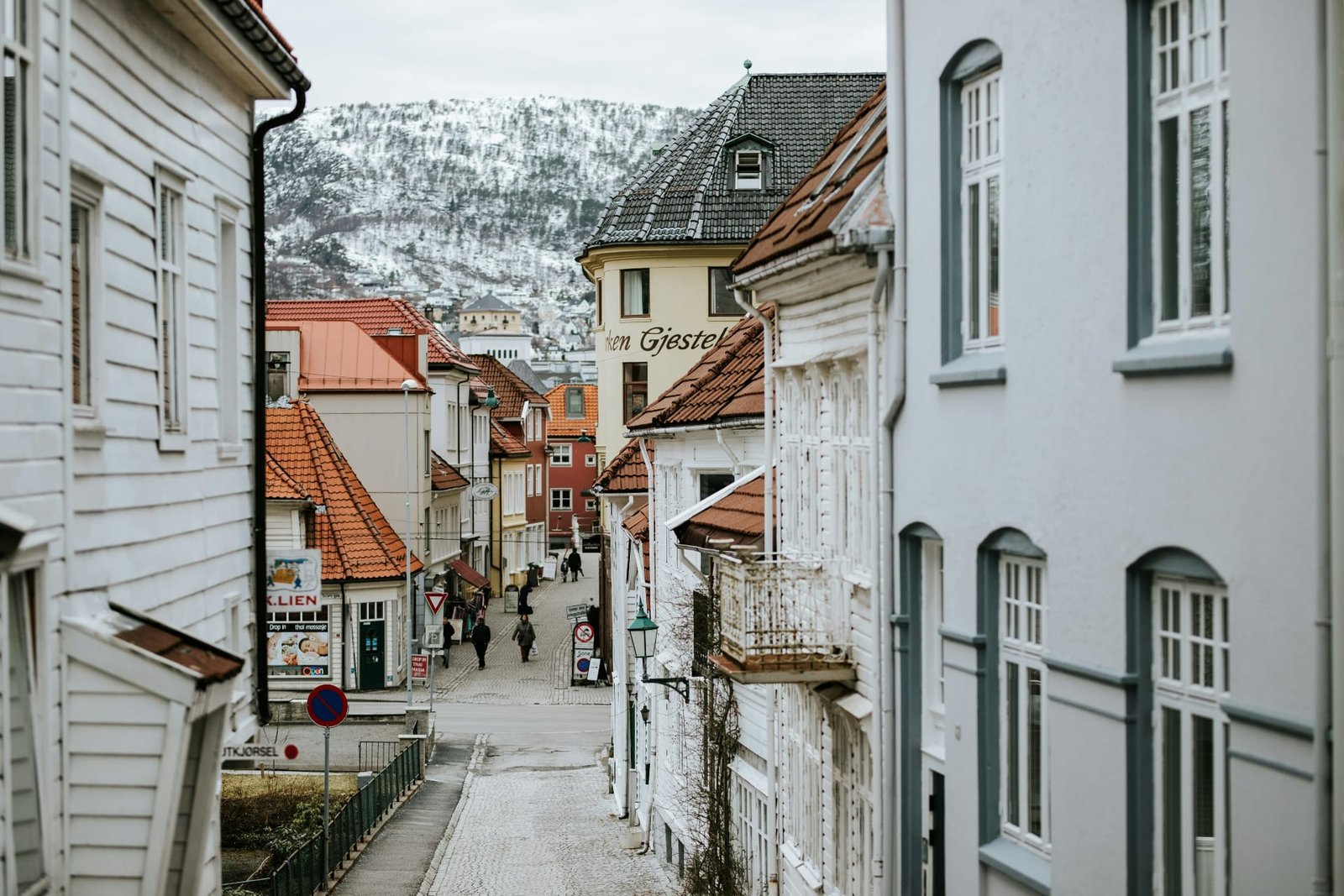 Charming narrow street in Bergen, Norway, lined with traditional wooden houses and red rooftops against a backdrop of snowy mountains.